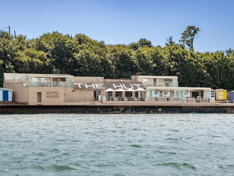 A photo of a waterside restaurant from the sea. It has the name 'The Hut' on the awning and there are beach huts and trees visible