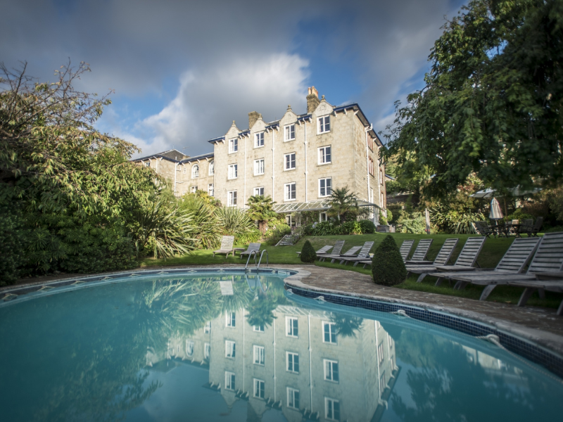 An imposing white hotel with a swimming pool and gardens in the foreground