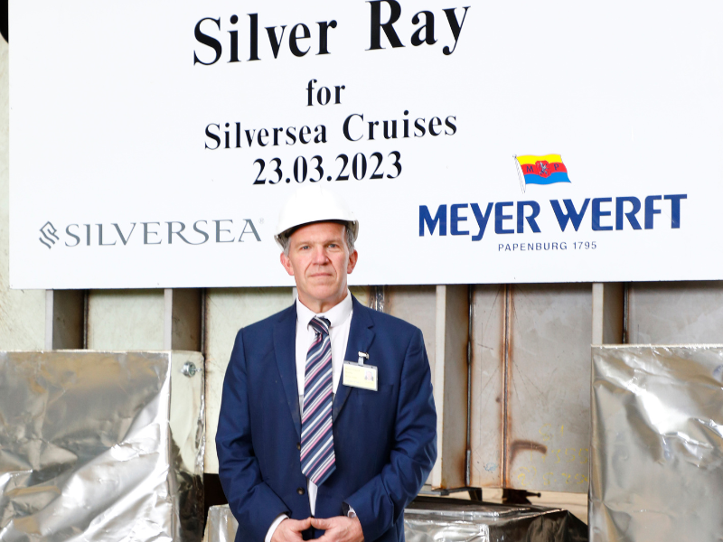 A man in a blue suit and white hard had stands in front of a sign saying 'Silver Ray for Silversea Cruises 28.03.23' - it's inside a shipyard in Germany
