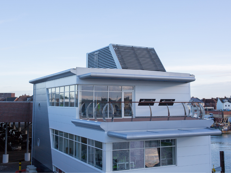A white building with lots of panoramic windows. The sea is visible behind