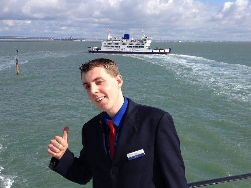 A young man wearing a suit has his thumbs up and is posing in front of a Wightlink ferry on the sea