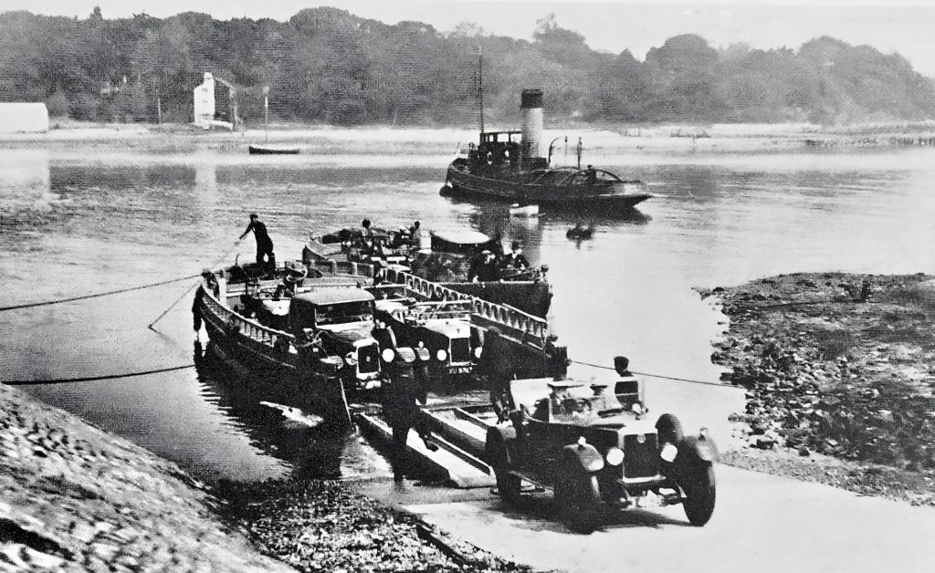 Black and white photo of wooden tow-boats carrying vintage cars being pulled across the water by a tug at Fishbourne in 1926.
