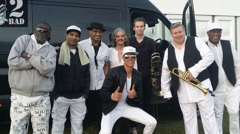 Eight members of reggae tribute band Johnny2Bad posing together in front of a tour van, with brass instruments and casual stage outfits ahead of a live performance.