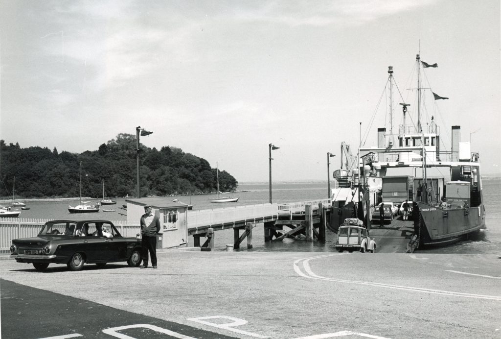 Black and white photograph of MV Camber Queen loading cars at a slipway, with vehicles driving on board and a crew member standing beside a parked car near the pier.