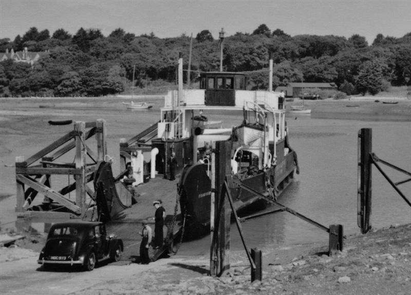 Black and white image of MV Fishbourne loading a car via hinged bow ramps at a slipway, with crew assisting at the water’s edge.