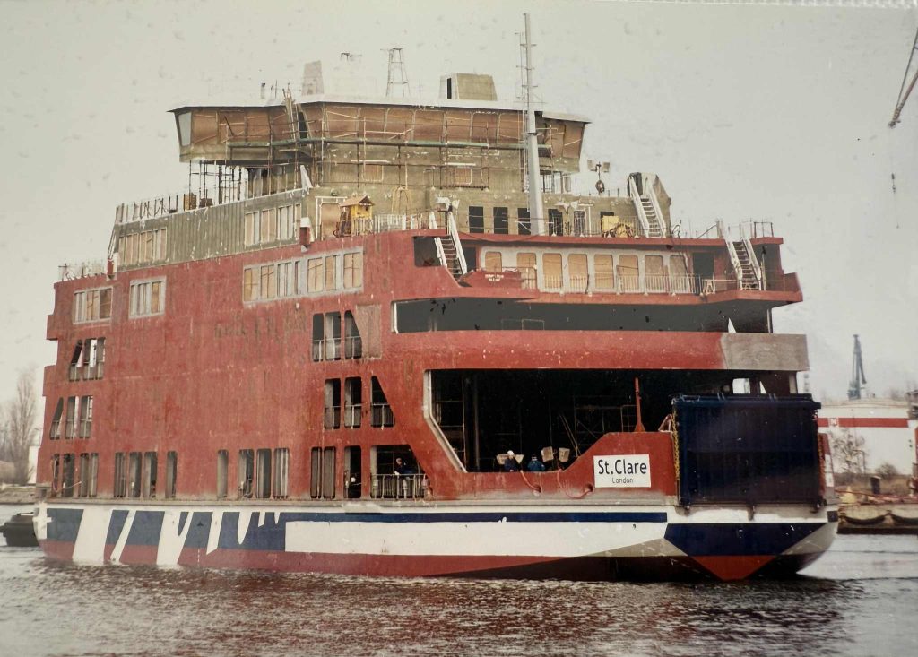MV St Clare afloat on the water during construction, showing the red hull and unfinished upper decks with scaffolding in place.