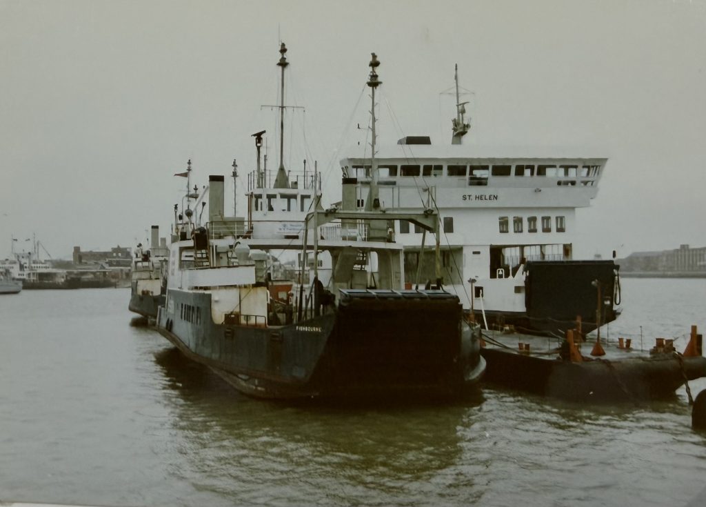 Historic ferries St Helen and Fishbourne moored side by side in harbour, viewed from the stern with loading ramps raised and city buildings in the background.