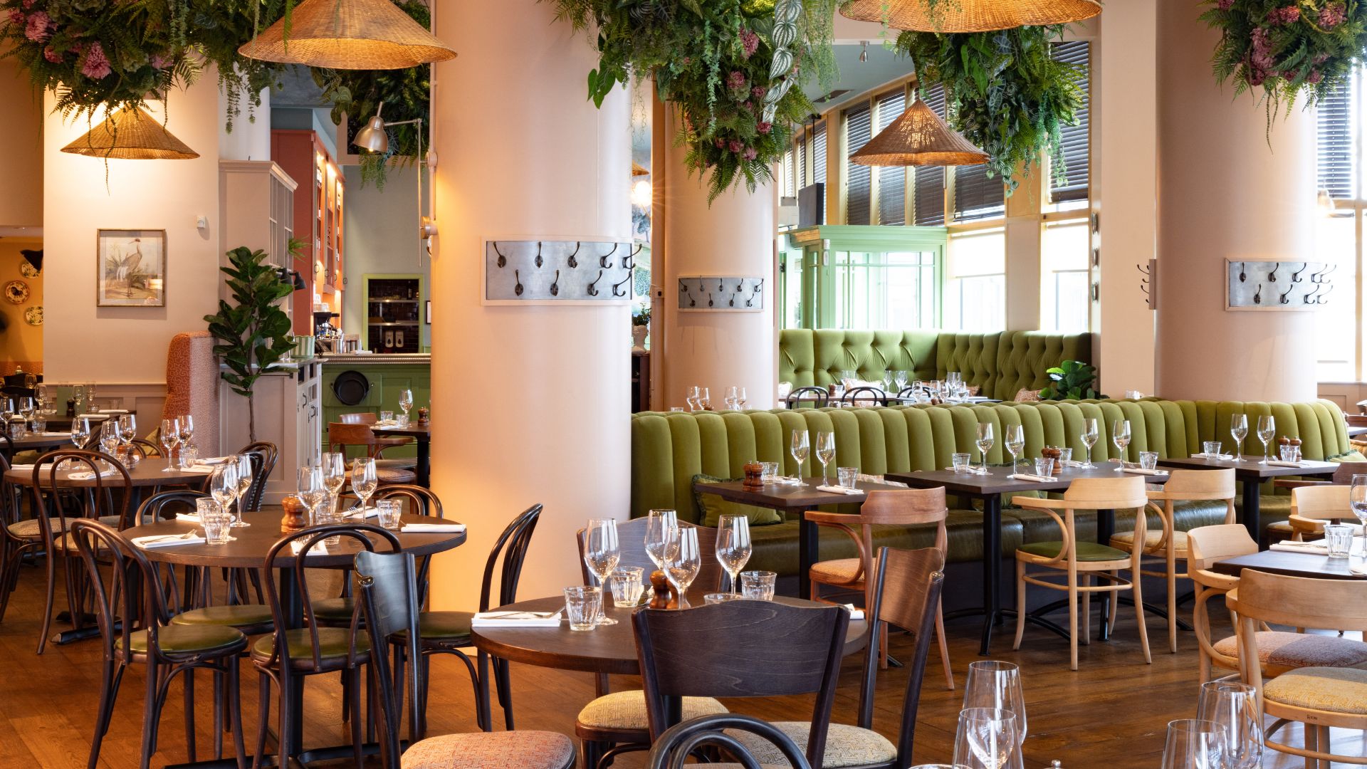 Elegant restaurant interior at Brasserie Blanc with wooden tables, green banquette seating and hanging plants.