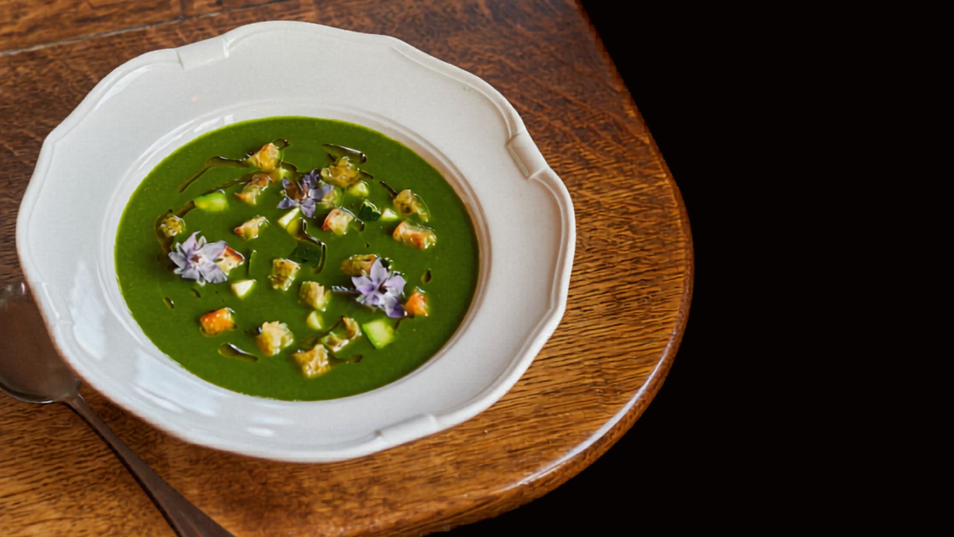 Bowl of green soup at Brasserie Blanc topped with croutons, herbs and edible flowers on a wooden table.