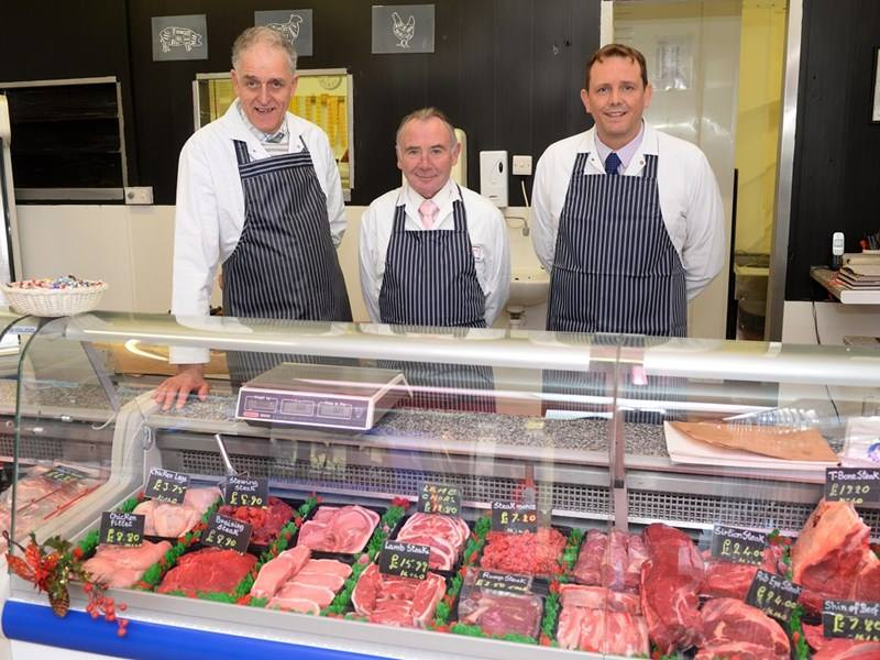 Three smiling butchers wearing blue and white striped aprons stand behind a meat counter at a butcher on the Isle of Wight