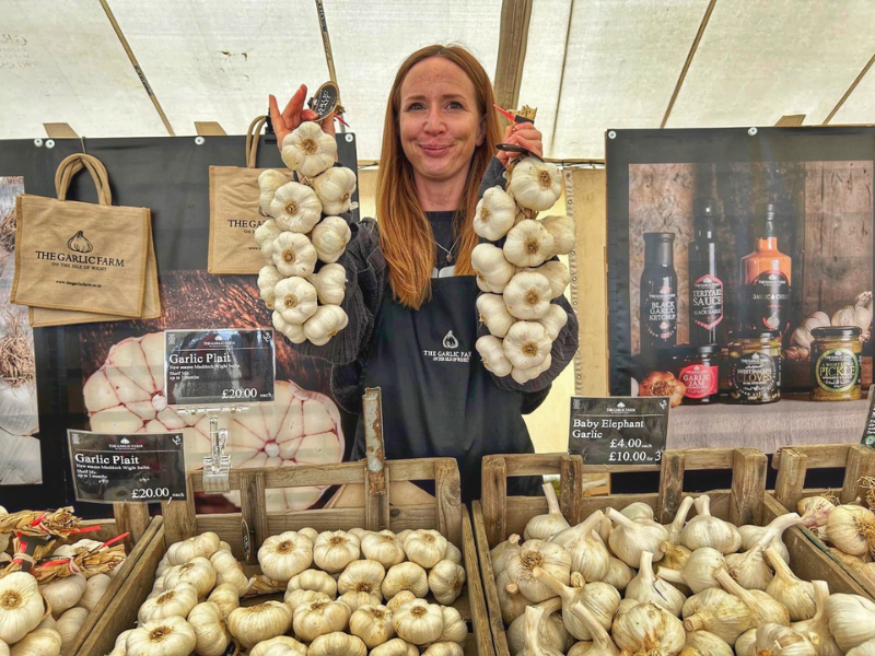 A woman holding up two plaits of garlic, in front of a garlic stall with lots of garlic displayed