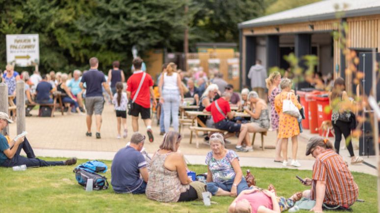 Visitors relaxing on grass and benches at an outdoor festival with food stalls and seating area