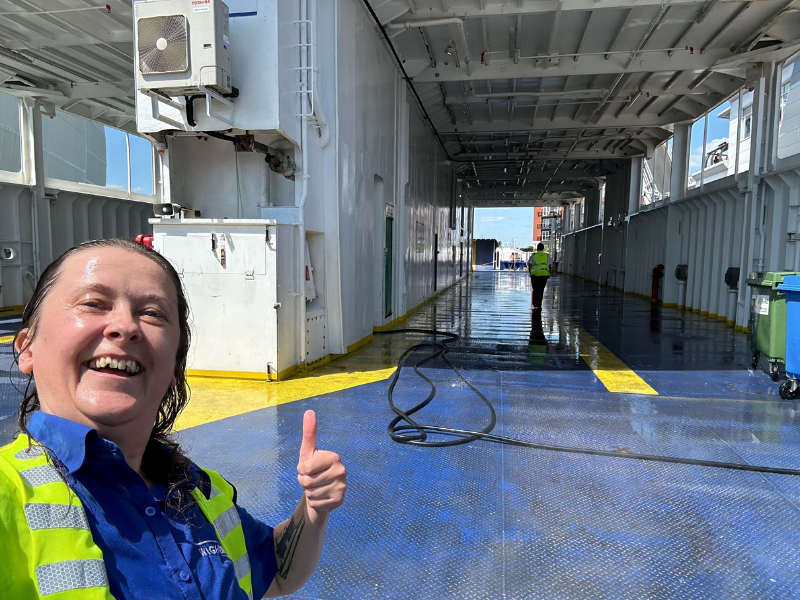 A woman with a blue uniform and yellow high visibility jacket has her thumbs up standing in a wet car deck of a ferry - the drencher system in operation