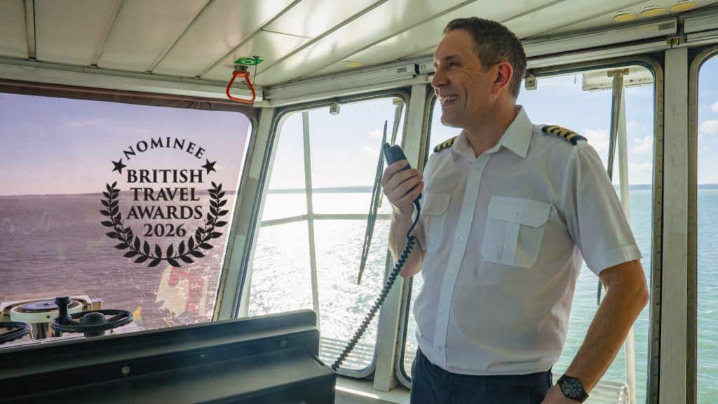 Ferry bridge interior with a Captain holding a handheld radio, large windows overlooking the sea, and a “British Travel Awards 2026 Nominee” badge displayed on the glass.