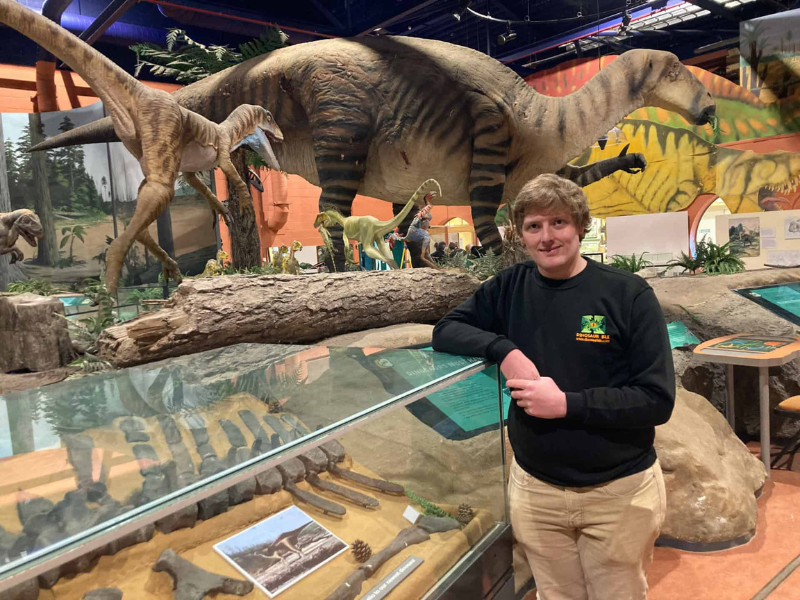 A man leaning against a museum exhibit in a glass case. There's a large replica dinosaur model in the background