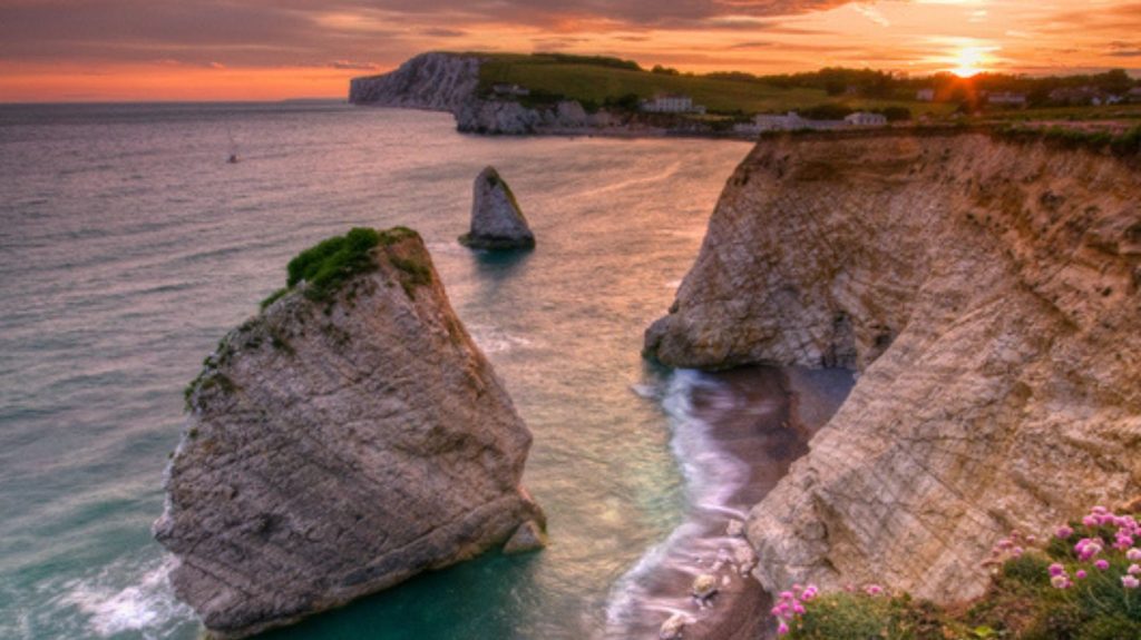 Sunset view over Freshwater Bay, with dramatic white chalk stacks and towering cliffs glowing in warm evening light above the sea.