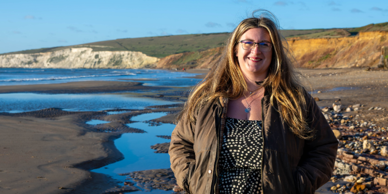 Author Megan Jacobs stands on a beach on the Isle of Wight, smiling for the camera. Image thanks to Clare Jacobs