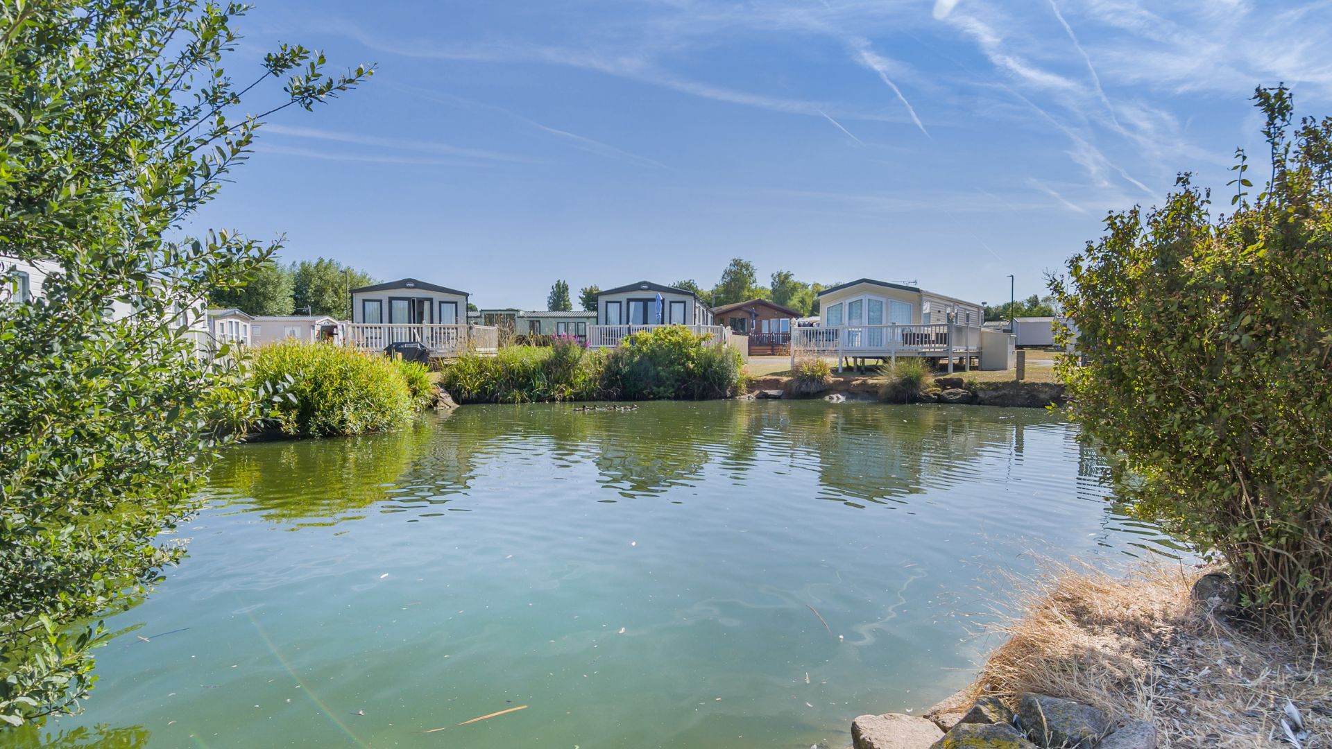 Holiday lodges overlooking a tranquil lake surrounded by trees and greenery at Manor Park holiday park.