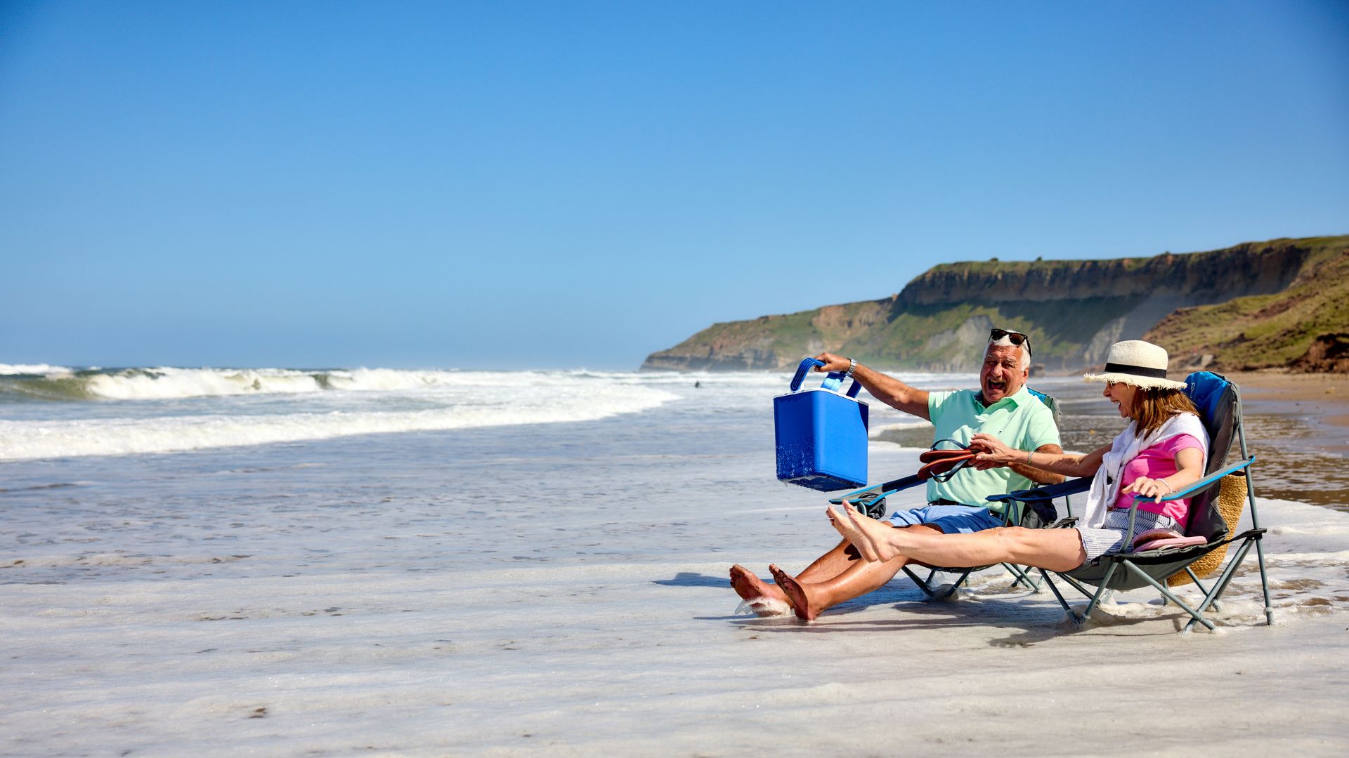 Couple sitting in beach chairs at the water’s edge, laughing as the waves wash around their feet on a sunny day.