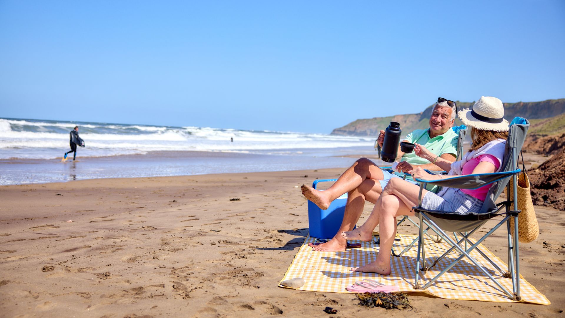 Couple relaxing on beach chairs beside a picnic blanket, enjoying hot drinks with surfers and cliffs in the distance.