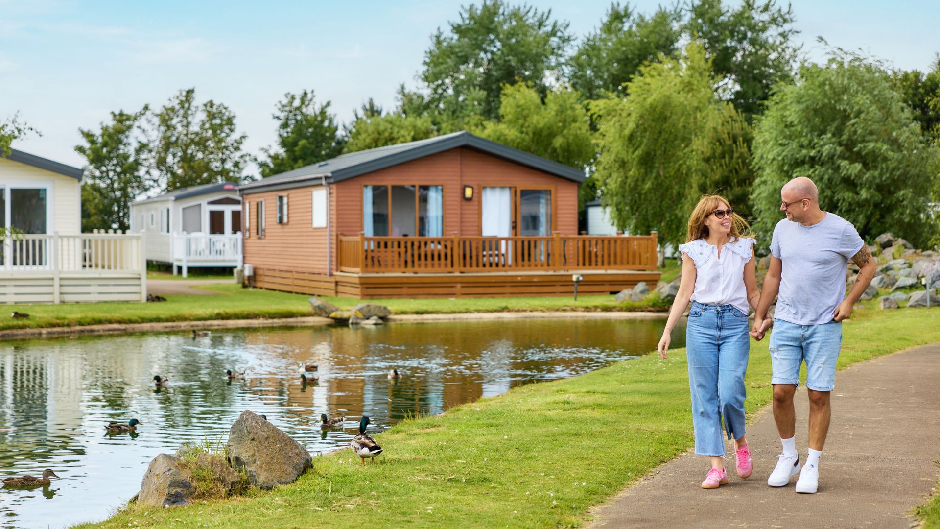 Couple holding hands and walking beside a lake, with modern holiday lodges and ducks in a peaceful park setting.