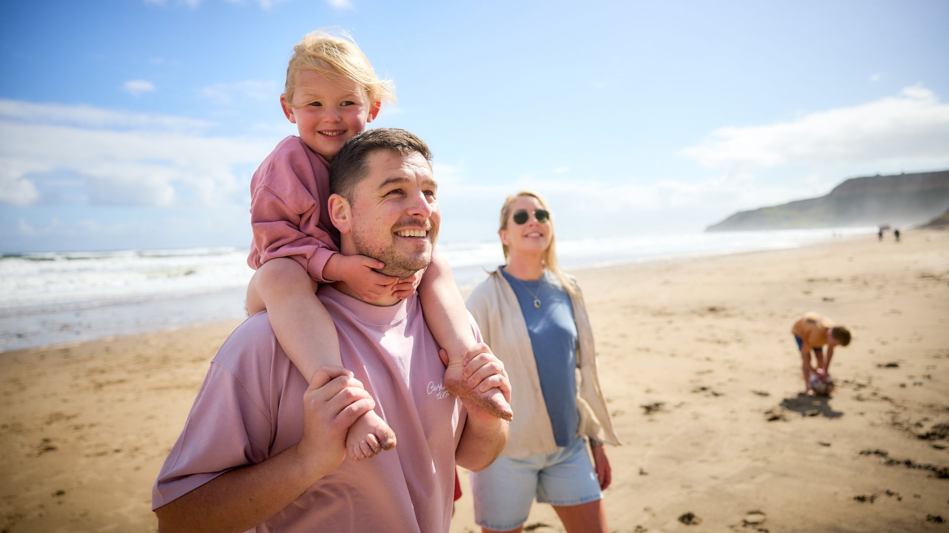 Young child sitting on their father's shoulders as a family enjoys a sunny walk along a sandy beach.