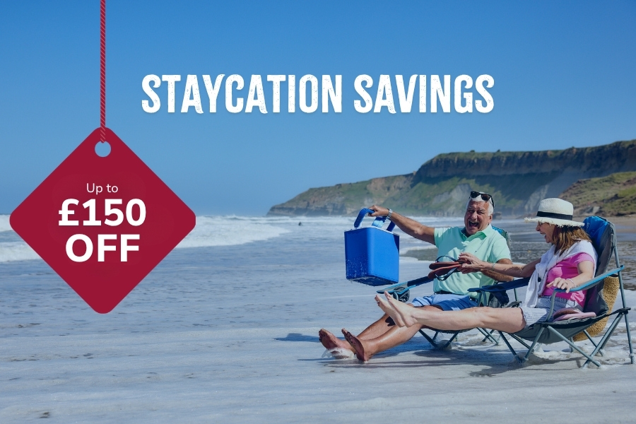 Promotional banner showing two adults relaxing on folding beach chairs by the sea with a red tag reading “Up to £150 off” and headline text “Staycation Savings”.