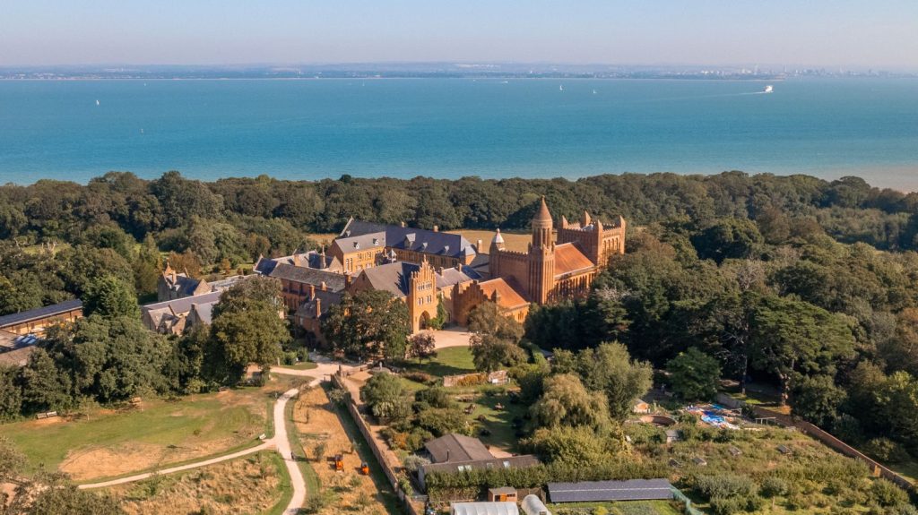 Aerial view of Quarr Abbey surrounded by woodland and gardens, with the Solent visible beyond the historic red-brick buildings.