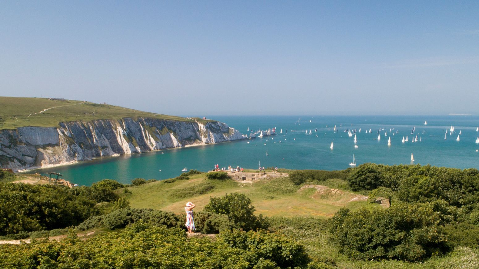 Panoramic view across the Needles headland, with white chalk cliffs, turquoise sea and dozens of sailing boats scattered across the Solent.