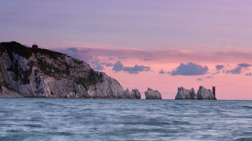 The Needles chalk stacks and lighthouse stand off the Isle of Wight coastline beneath a pink and purple sunset sky