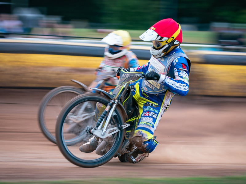 Two speedway bikes speeding around a track on the Isle of Wight, one of them is from the Wightlink Warriors