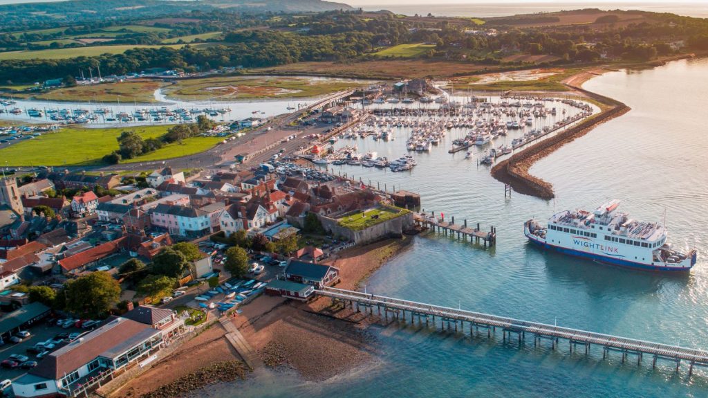Aerial view of Yarmouth Harbour with a Wightlink ferry arriving alongside the historic town, marina full of yachts and surrounding coastline in warm evening light.