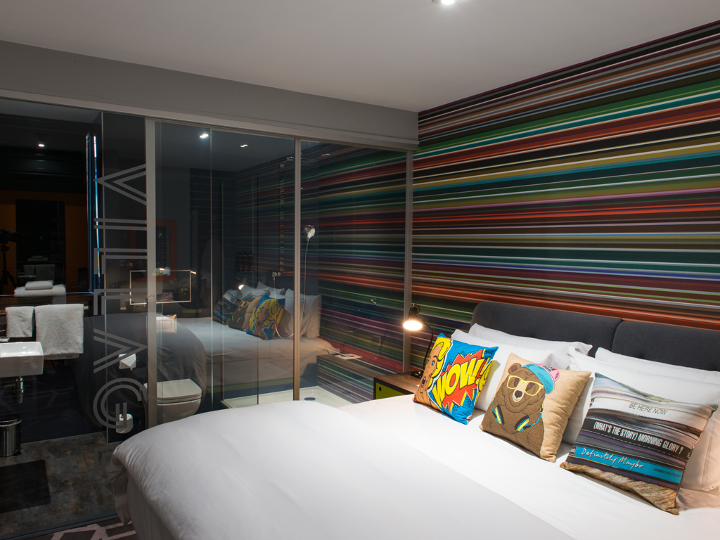 Modern Village Hotel bedroom with colourful striped feature wall, double bed and glass‑walled bathroom