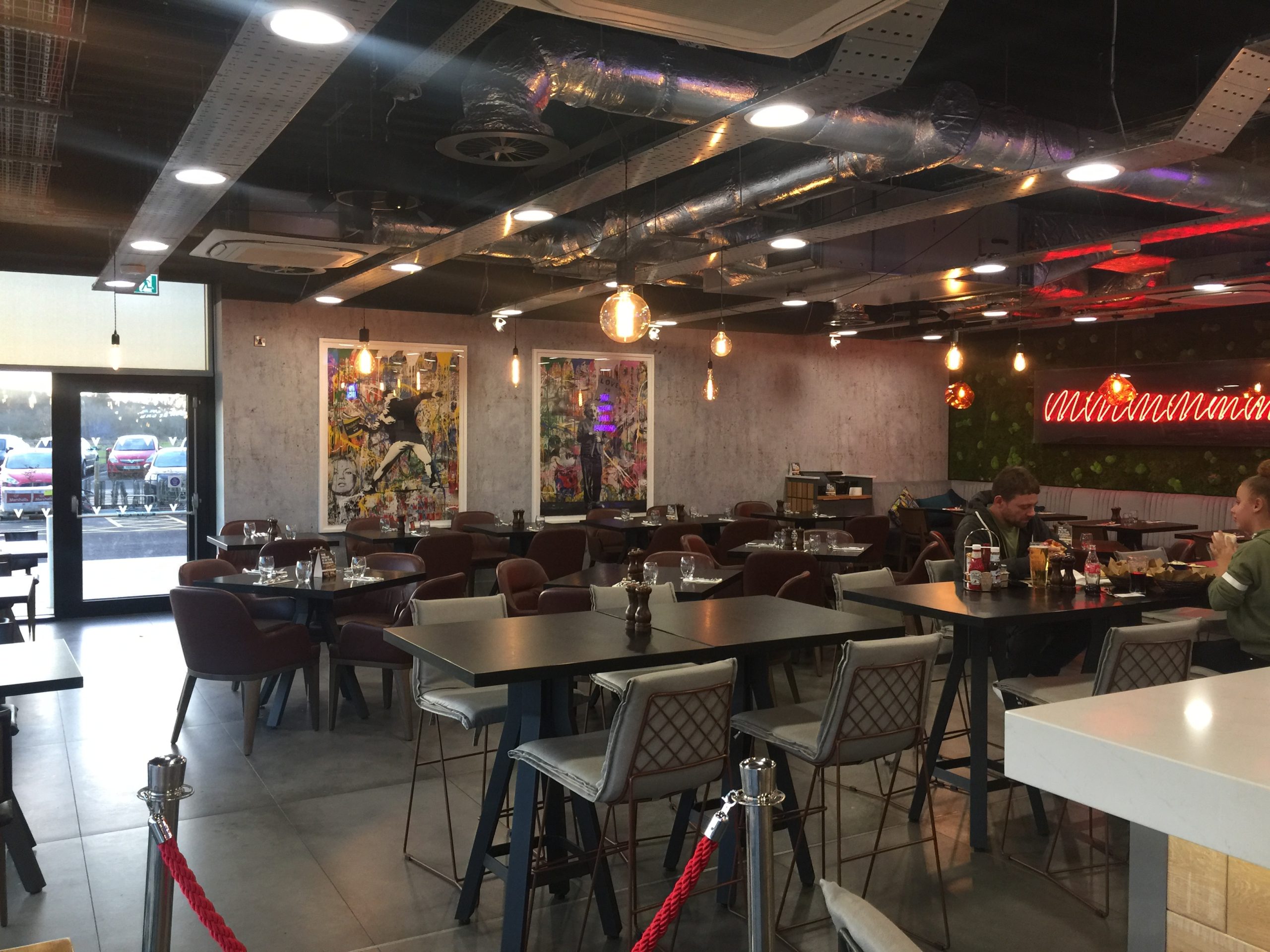 Village Hotel restaurant interior with modern tables, hanging lights, wall art and “V bar & grill” neon sign