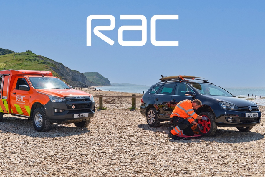 RAC roadside assistance van and patrol changing a car tyre on a pebble beach, with surfboards on the vehicle and coastline in the background.