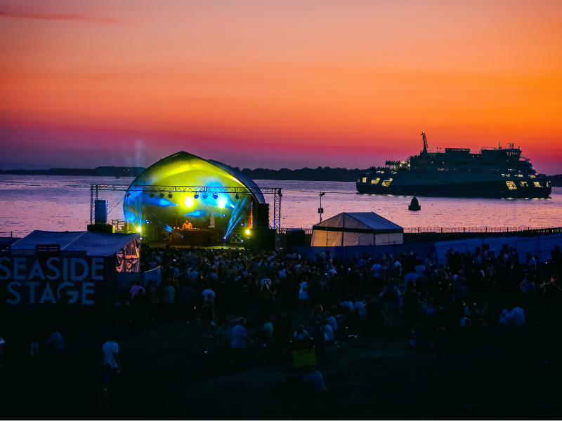 A music festival by the sea, pictured at Sunset, with a lit up stage, crowds and a ferry passing
