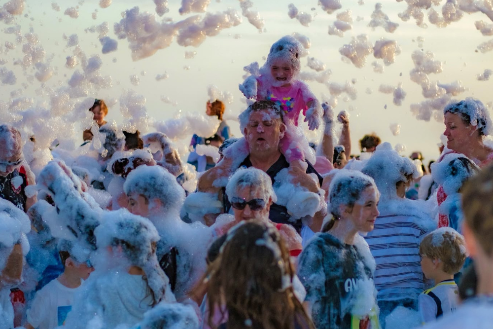 A large crowd of adults and children taking part in an outdoor foam party at sunset. Foam fills the air and covers participants, while one child sits on an adult's shoulders in the centre of the crowd.
