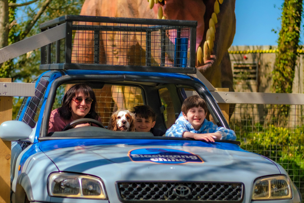Two children, an adult, and a dog sitting inside a blue safari-style vehicle at a dinosaur-themed attraction. A large dinosaur model is visible behind the vehicle within a fenced area.