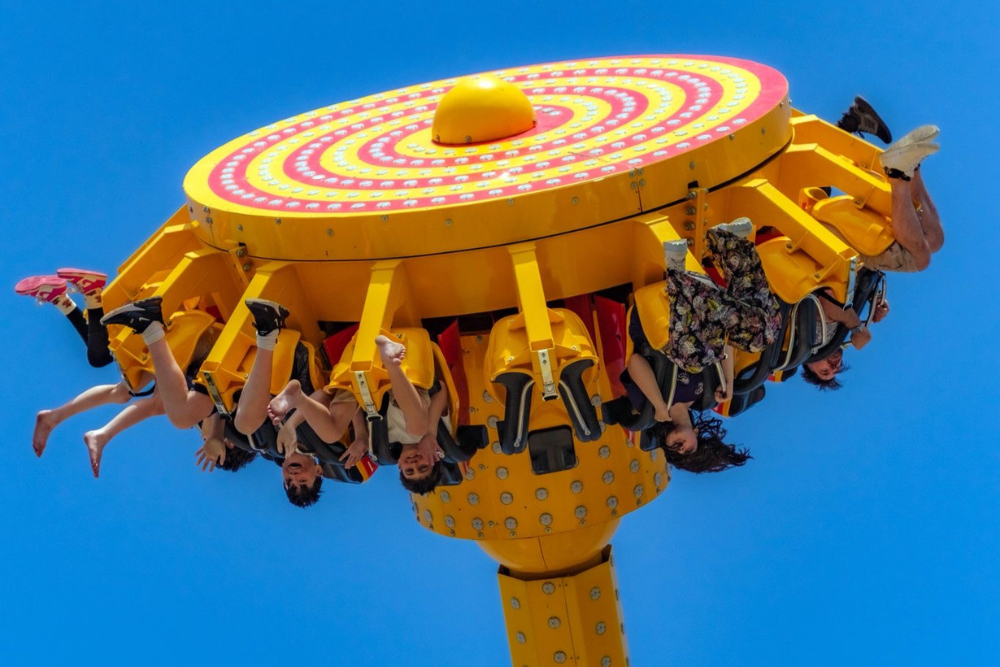 Several riders seated on a bright yellow amusement ride suspended upside down against a clear blue sky, with legs extended outward as the ride rotates.