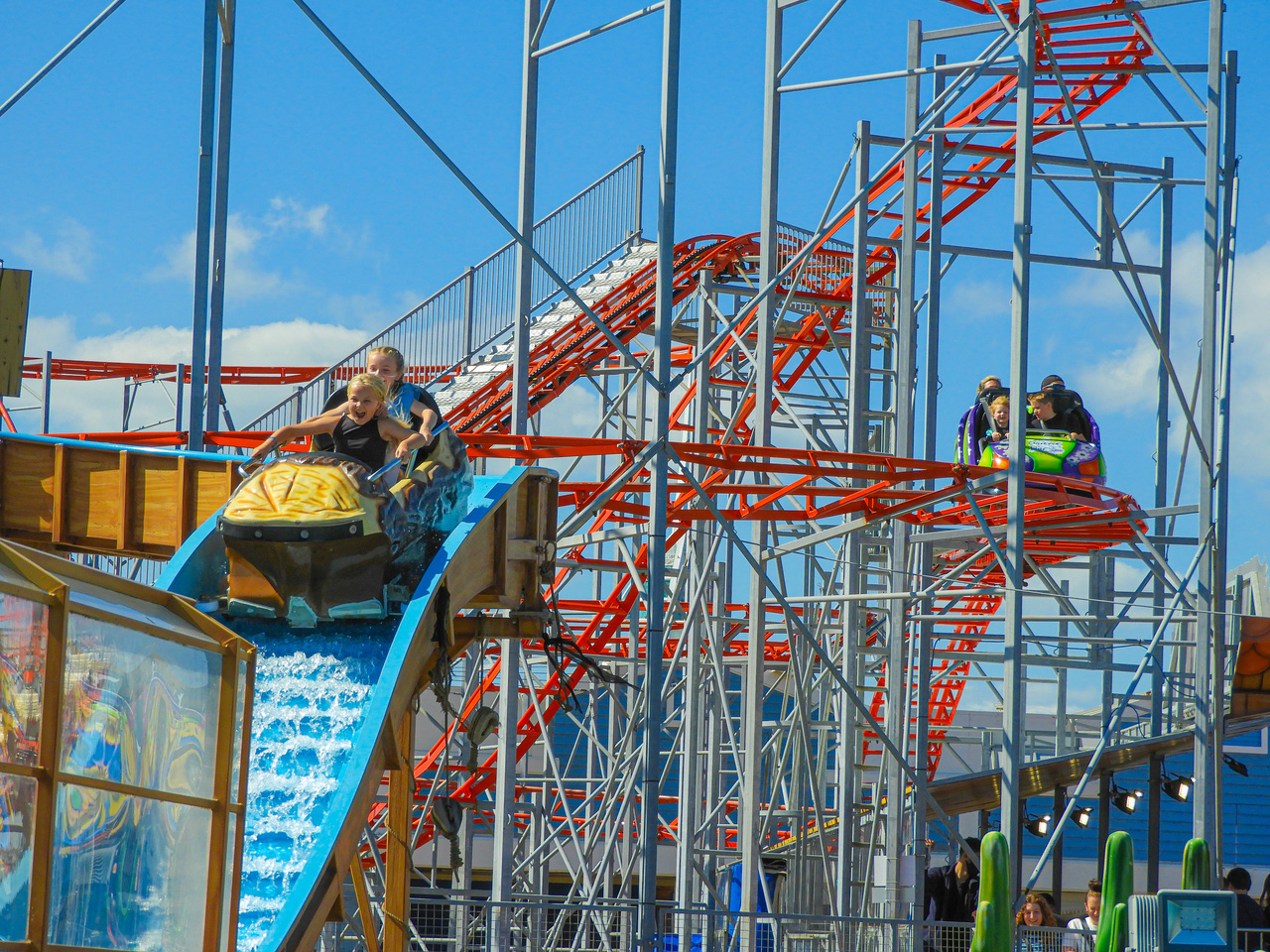 Visitors ride a rollercoaster through a network of bright red tracks and silver support structures, with a splash section in the foreground and a clear blue sky overhead.