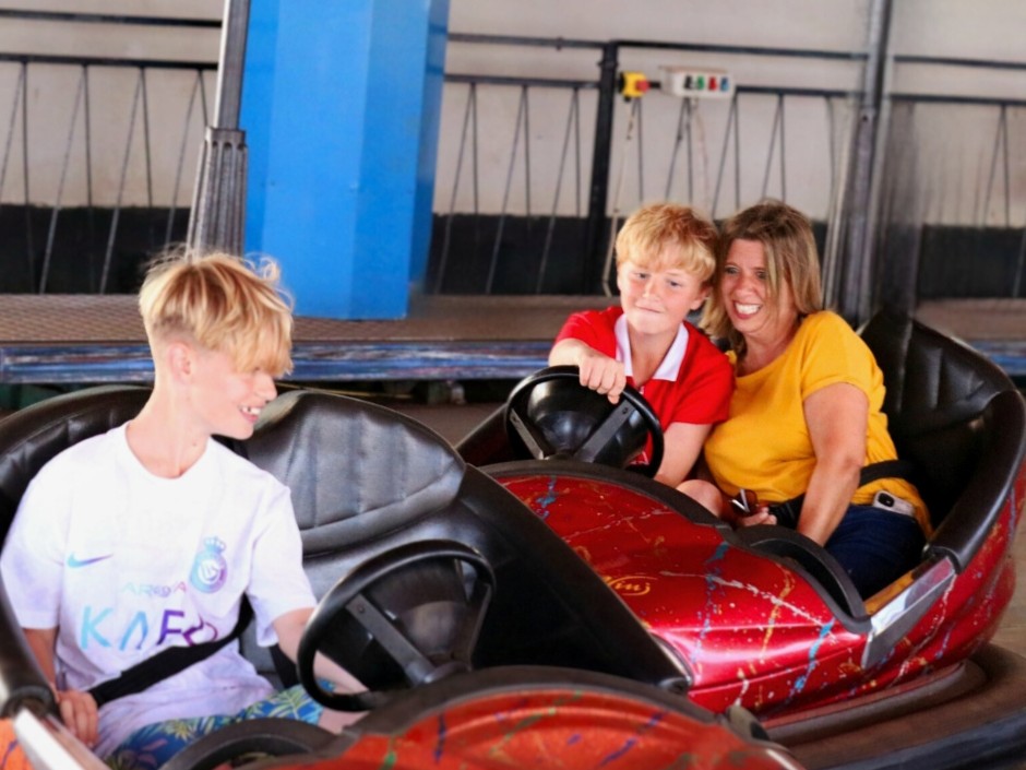 Three people riding bumper cars inside an amusement arcade, with two red cars positioned side by side on the dodgems track.