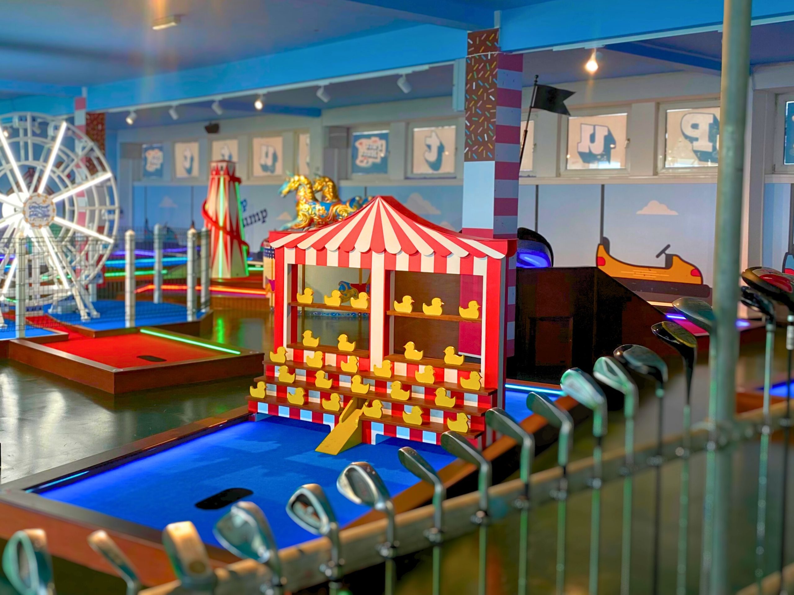 Indoor amusement area featuring a themed mini-golf course with golf clubs lined up beside a blue putting green, a striped carnival-style duck game, a ferris wheel attraction, and colourful neon lighting throughout.