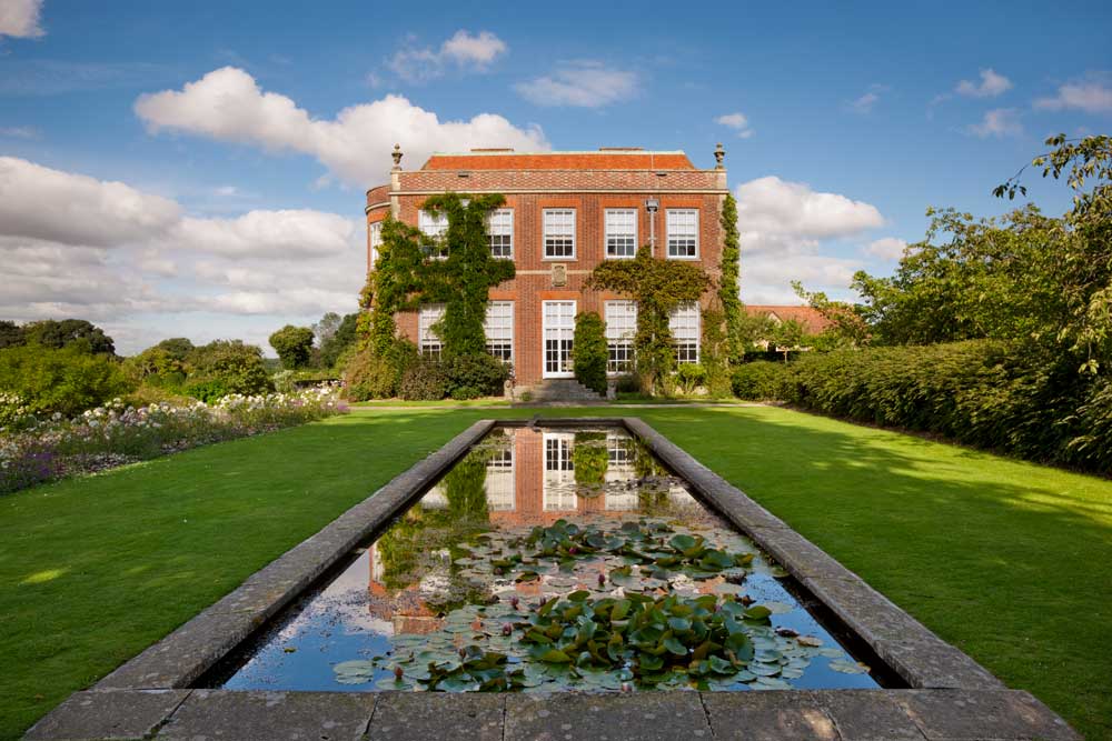 View of Hinton Ampner house from across a long lily pool surrounded by formal gardens under a blue sky.