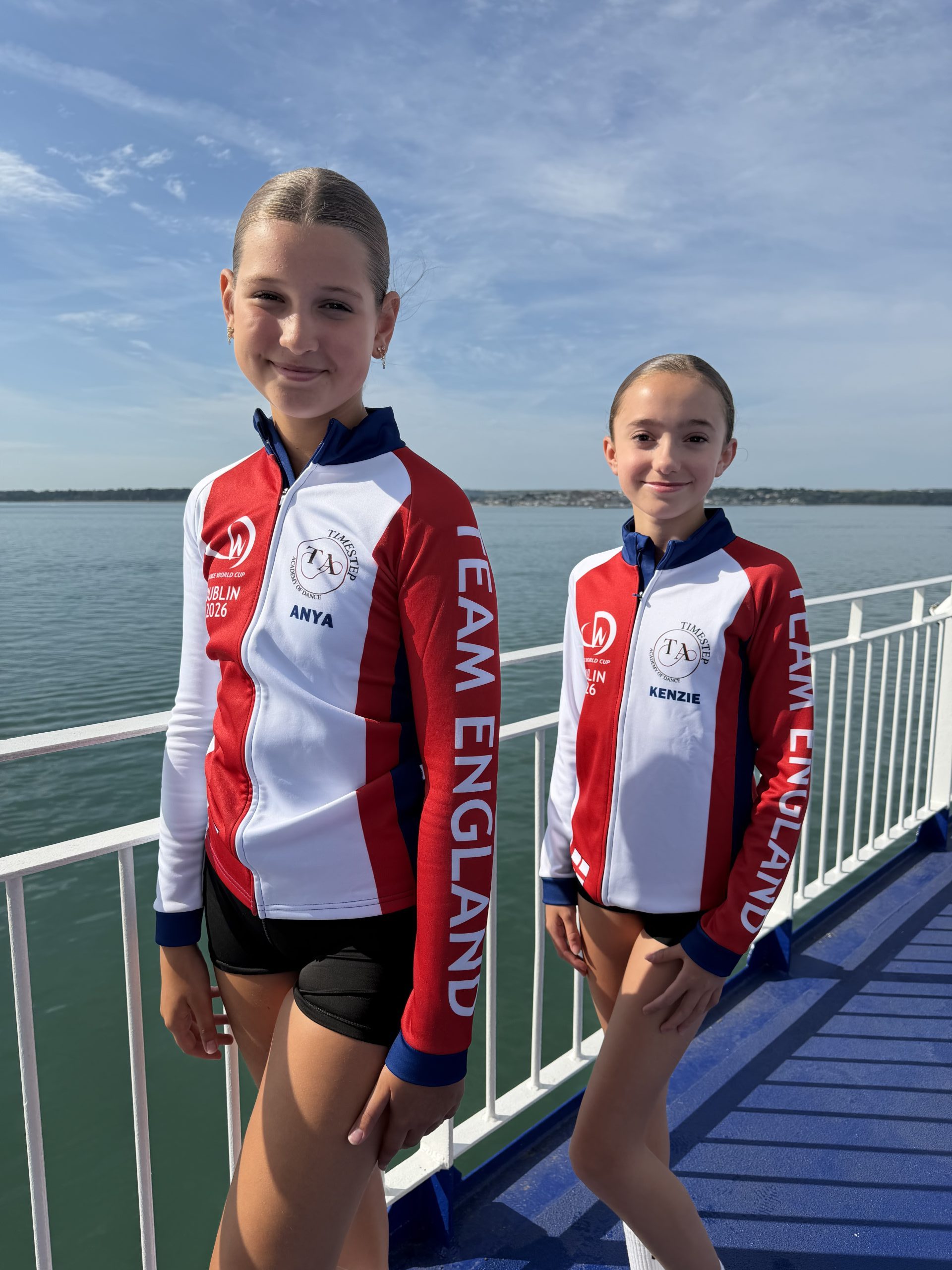 Two young Team England dancers in red, white and blue tracksuits stand on a ferry deck with the sea behind them, travelling to compete at the Dance World Cup in Dublin.