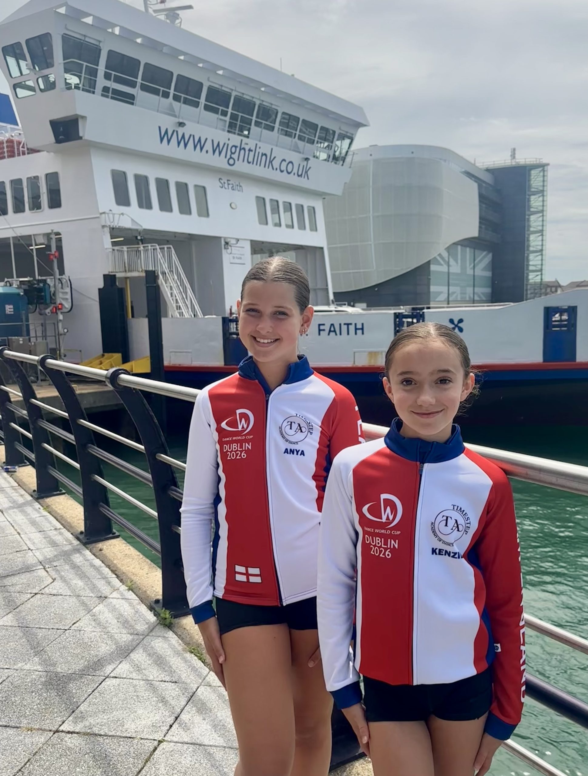 Two young dancers in Team England tracksuits pose in front of a Wightlink ferry, highlighting travel support to the Dance World Cup Finals in Dublin.