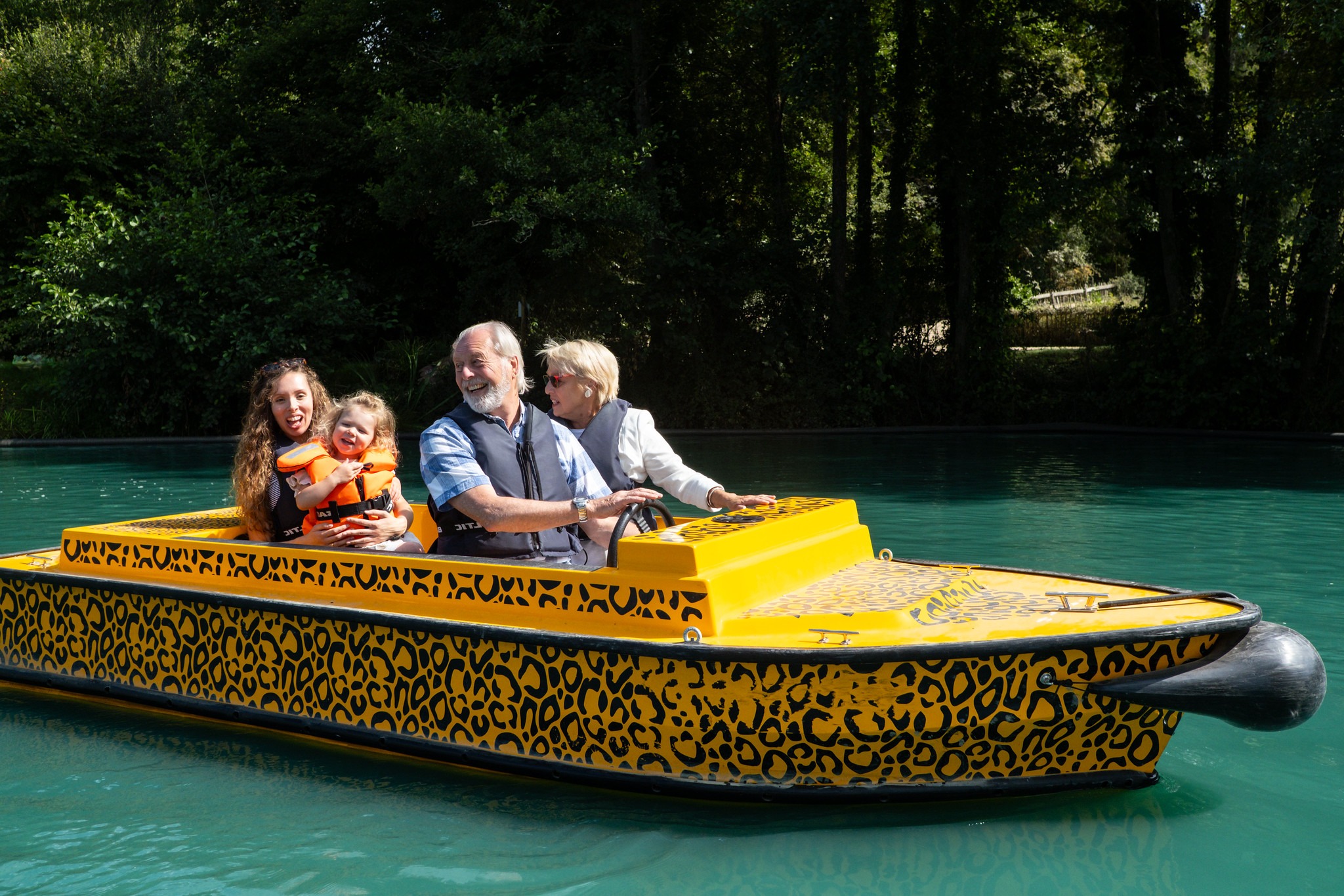 Four people wearing life jackets ride in a yellow, animal-print electric boat on a turquoise lagoon surrounded by trees.