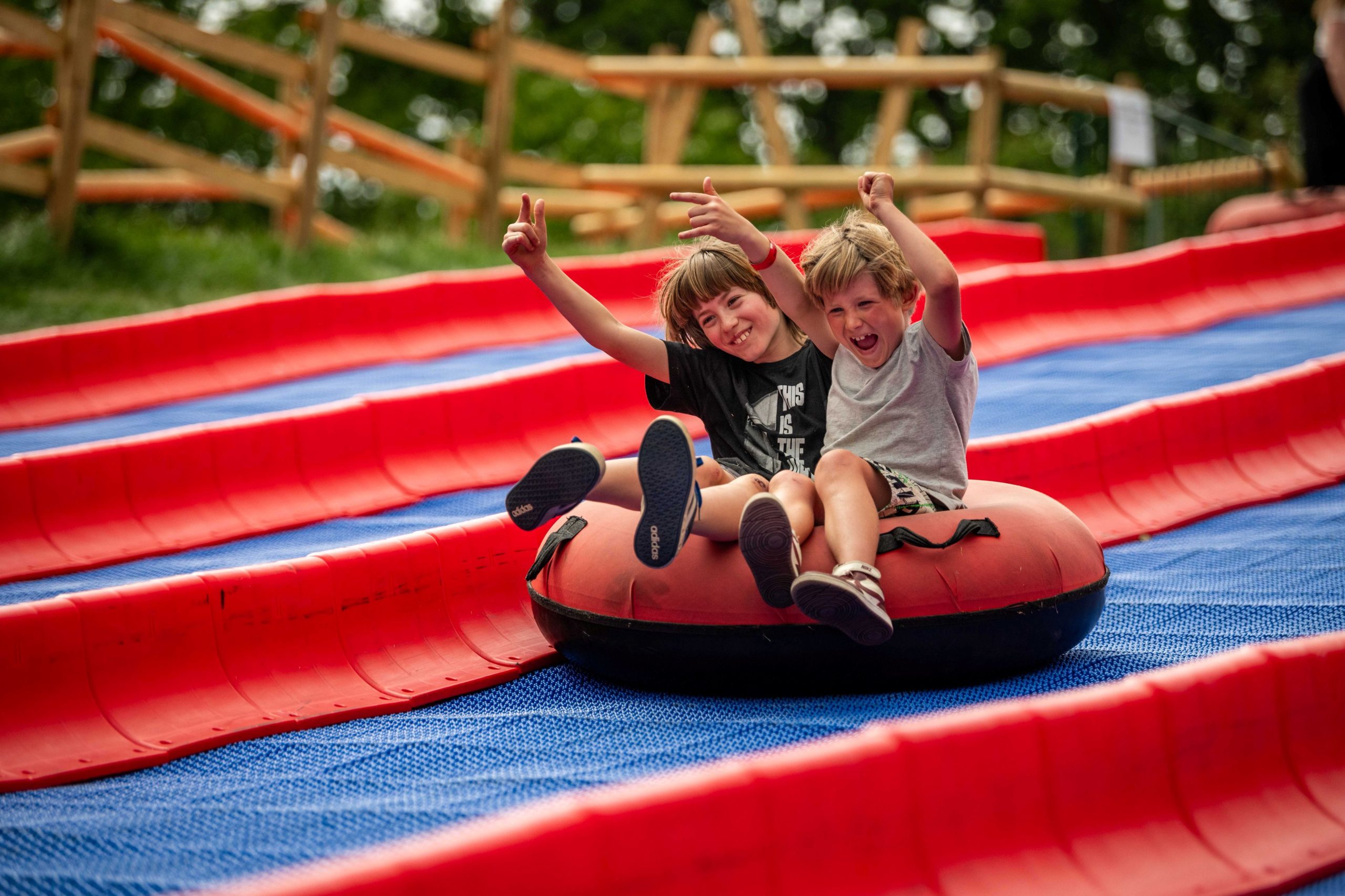 Two children ride together in an inflatable tube down a multi-lane outdoor tubing slide with bright red sides and a blue track.