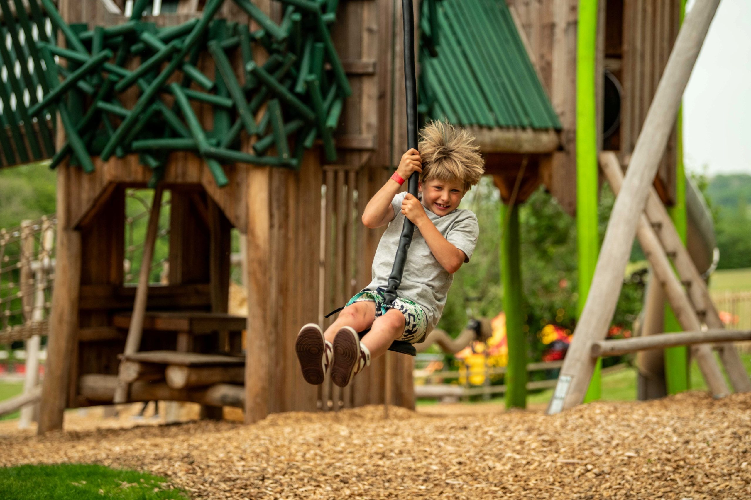 A child rides a zip line across a wooden adventure playground with climbing structures and woodchip ground cover.