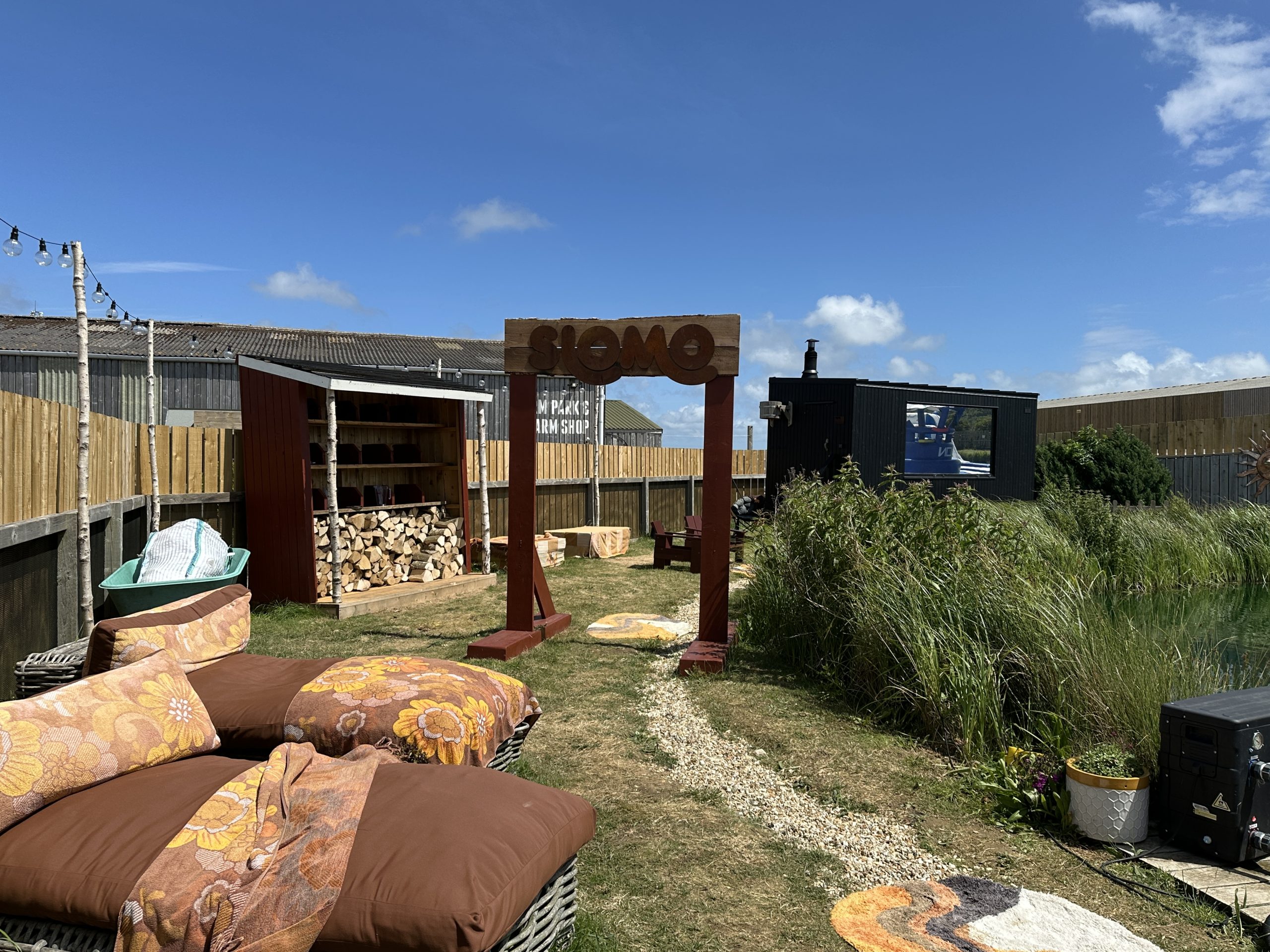 Outdoor seating area beside the SLOMO sauna at Tapnell Farm, overlooking a pond under clear blue skies on the Isle of Wight.