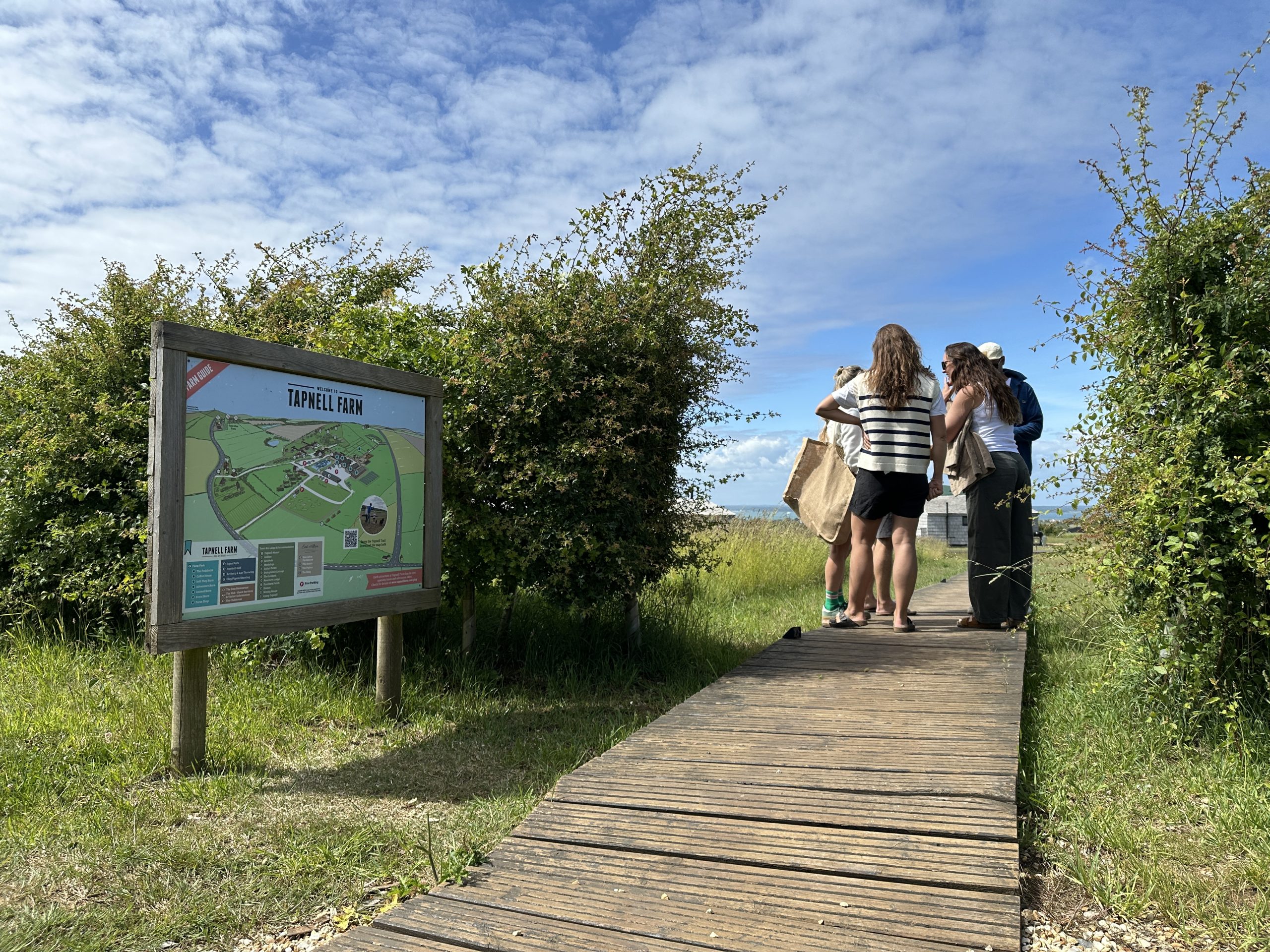 Visitors stand beside a Tapnell Farm map board on a wooden walkway surrounded by greenery and open countryside on the Isle of Wight.